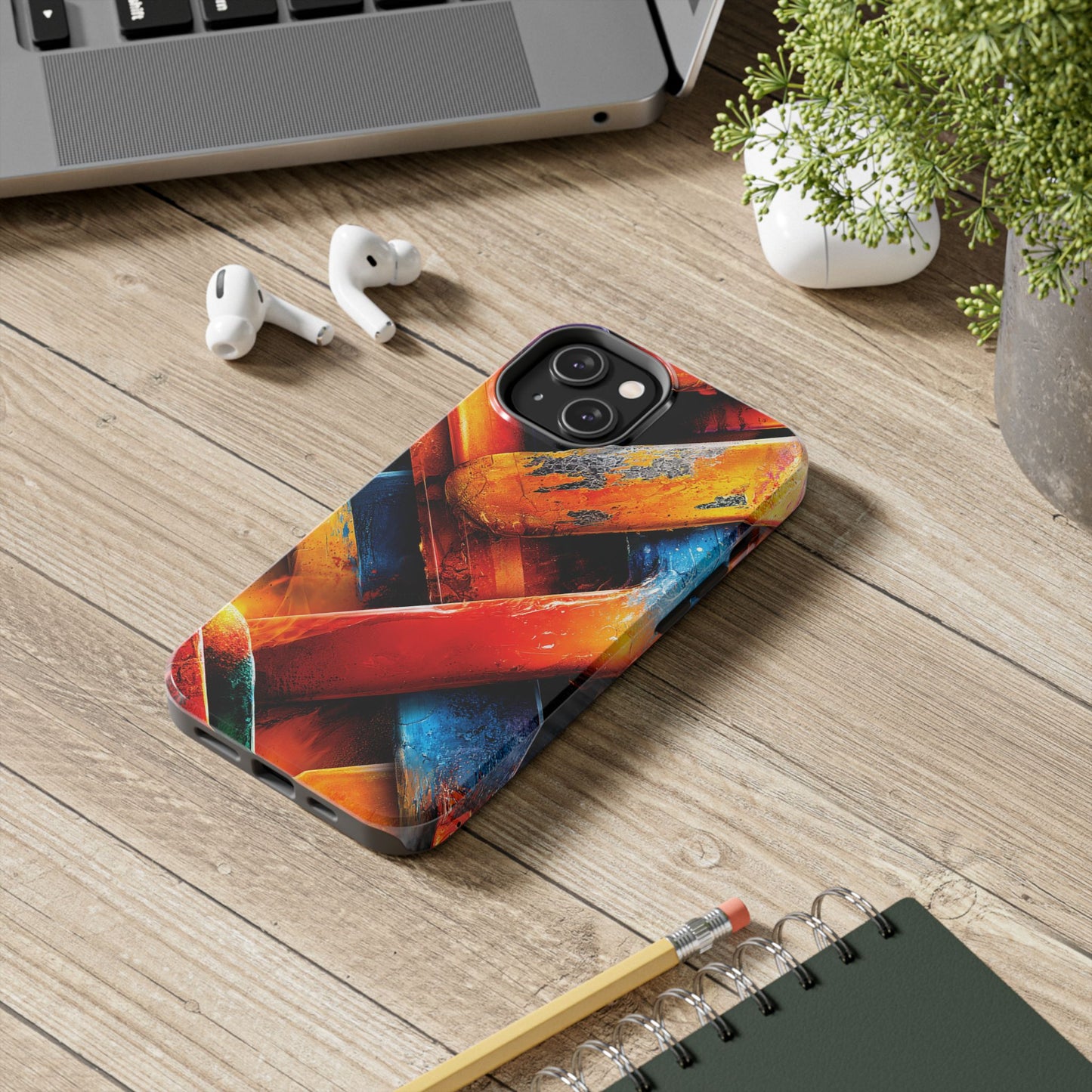 Colorful phone case on a wooden surface with a laptop, earbuds, and notebook.